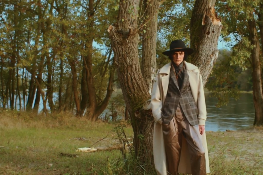 Persona in un cappotto bianco e cappello, appoggiata a un albero vicino a un lago.