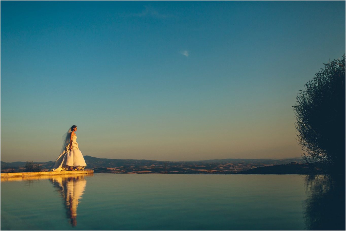 Sposa in abito bianco cammina lungo un bordo di piscina al tramonto.