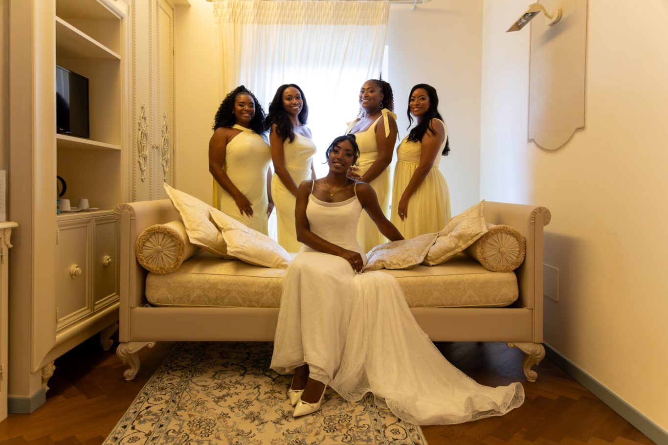 Sposa in abito bianco seduta su un divano, circondata da cinque damigelle in abiti gialli.