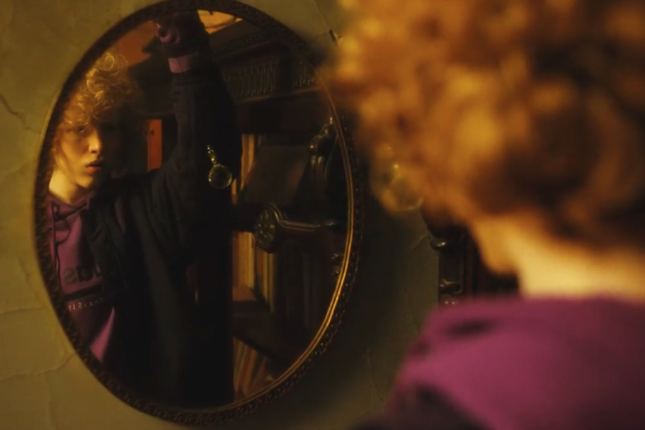 Riflesso di una persona con capelli ricci rossi in uno specchio, vestita di viola.