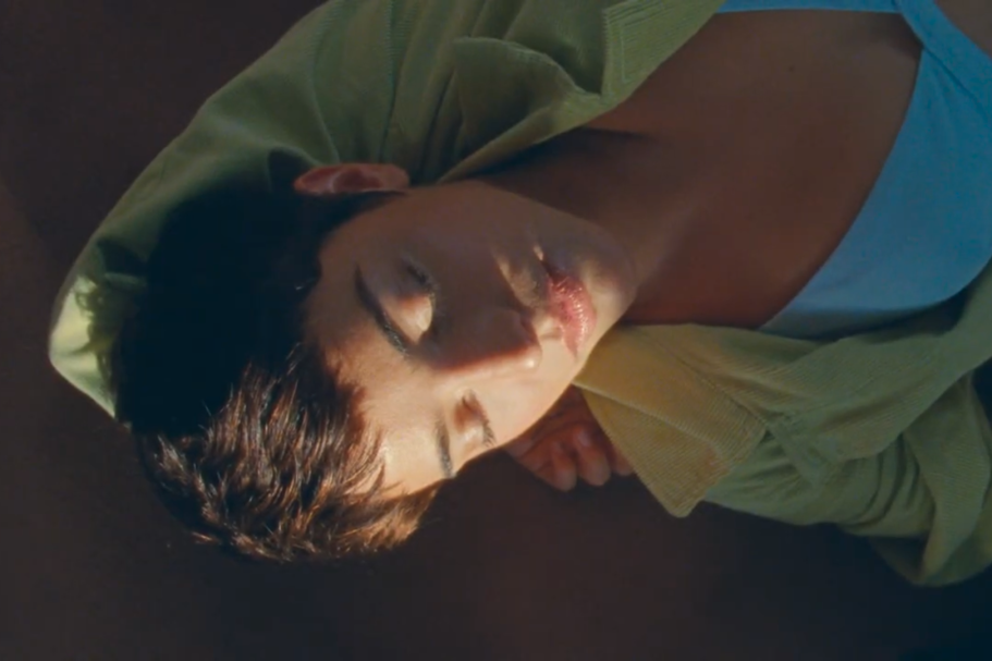 Ragazza sdraiata con camicia gialla, sguardo sereno e luce soffusa sul viso.