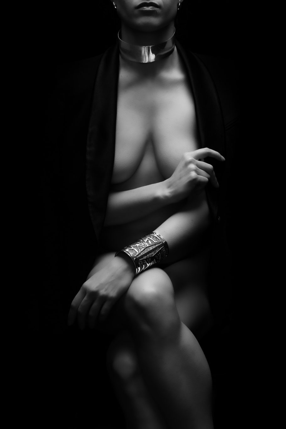 Figura nuda seduta, parzialmente drappeggiata, in condizioni di scarsa illuminazione con enfasi su mani e accessori.