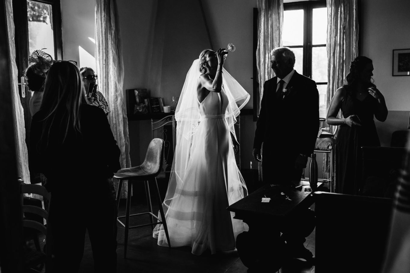 Sposa in abito bianco e velo, circondata da ospiti in una stanza scarsamente illuminata.