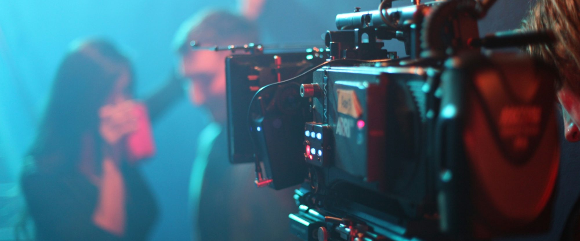 Una cinepresa con sfondo di una festa, persone che si divertono e bevono.