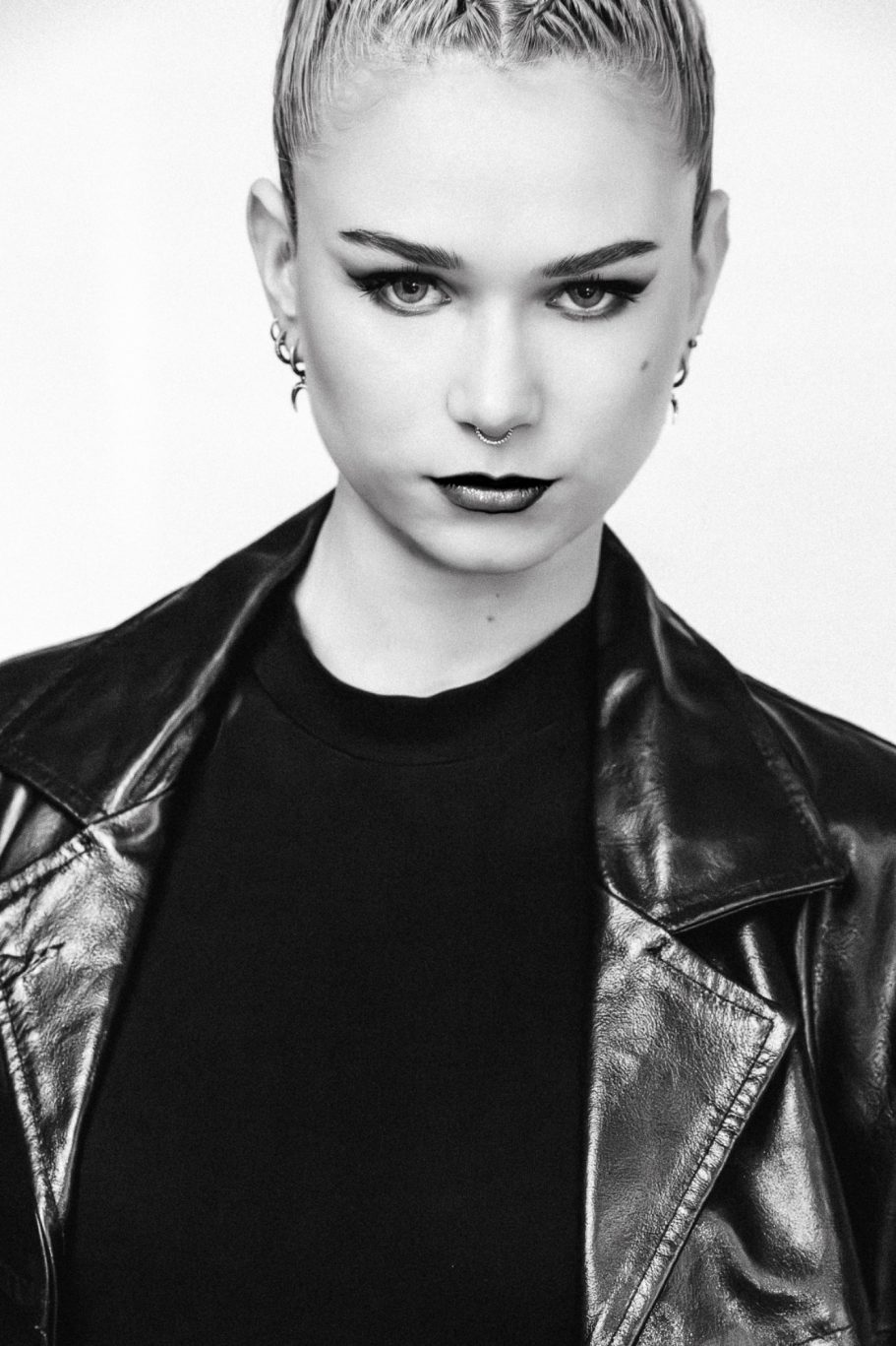 Ritratto in bianco e nero di una persona con capelli corti e trucco intenso, indossando un giubbotto di pelle.